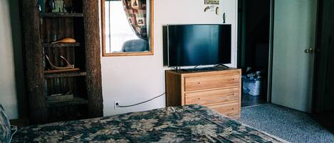 A bedroom with a bed, a dresser, a television, a mirror, and a shelf with various items.