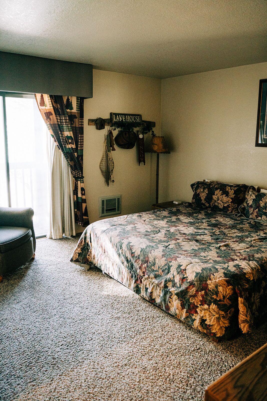 Une chambre à coucher avec une couvre-lit à motifs floraux, une chaise, un panneau indiquant « MAISON AU BORD DU LAC » et une fenêtre avec des rideaux.