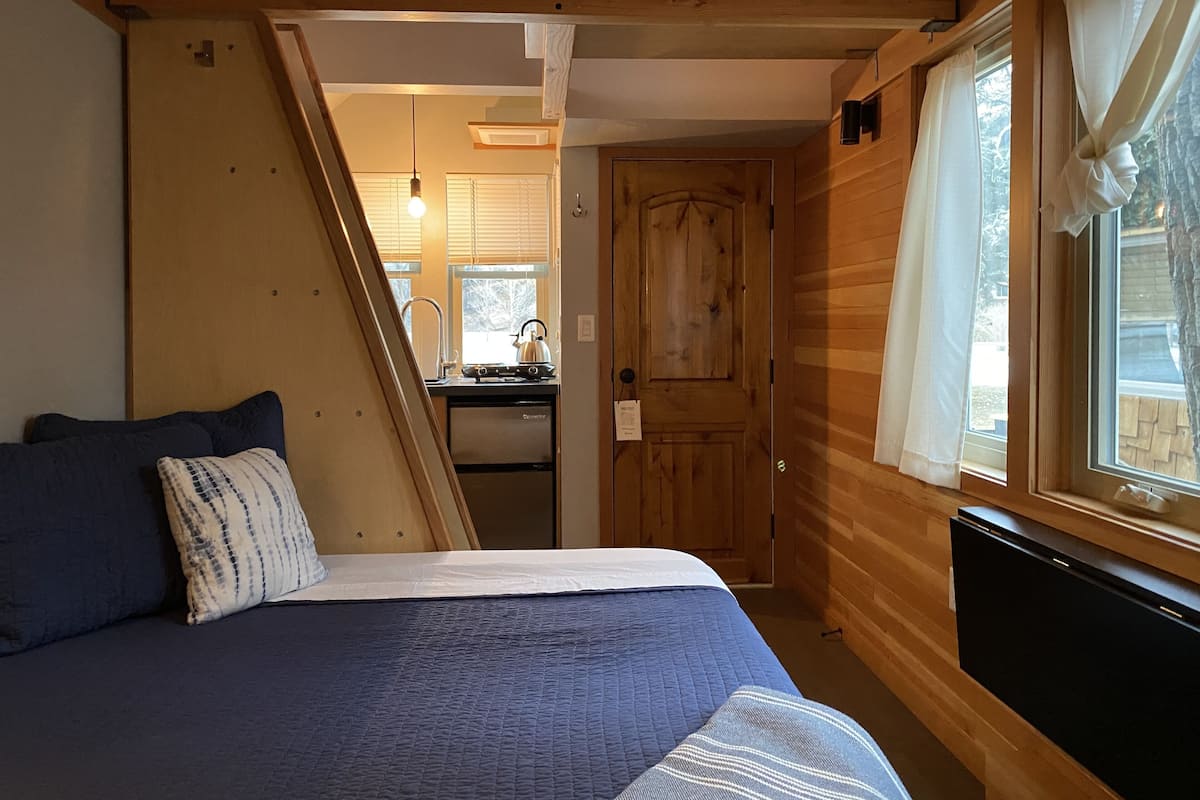 A compact bedroom with a bed, a window with curtains, a wooden door, and a kitchenette in the background.