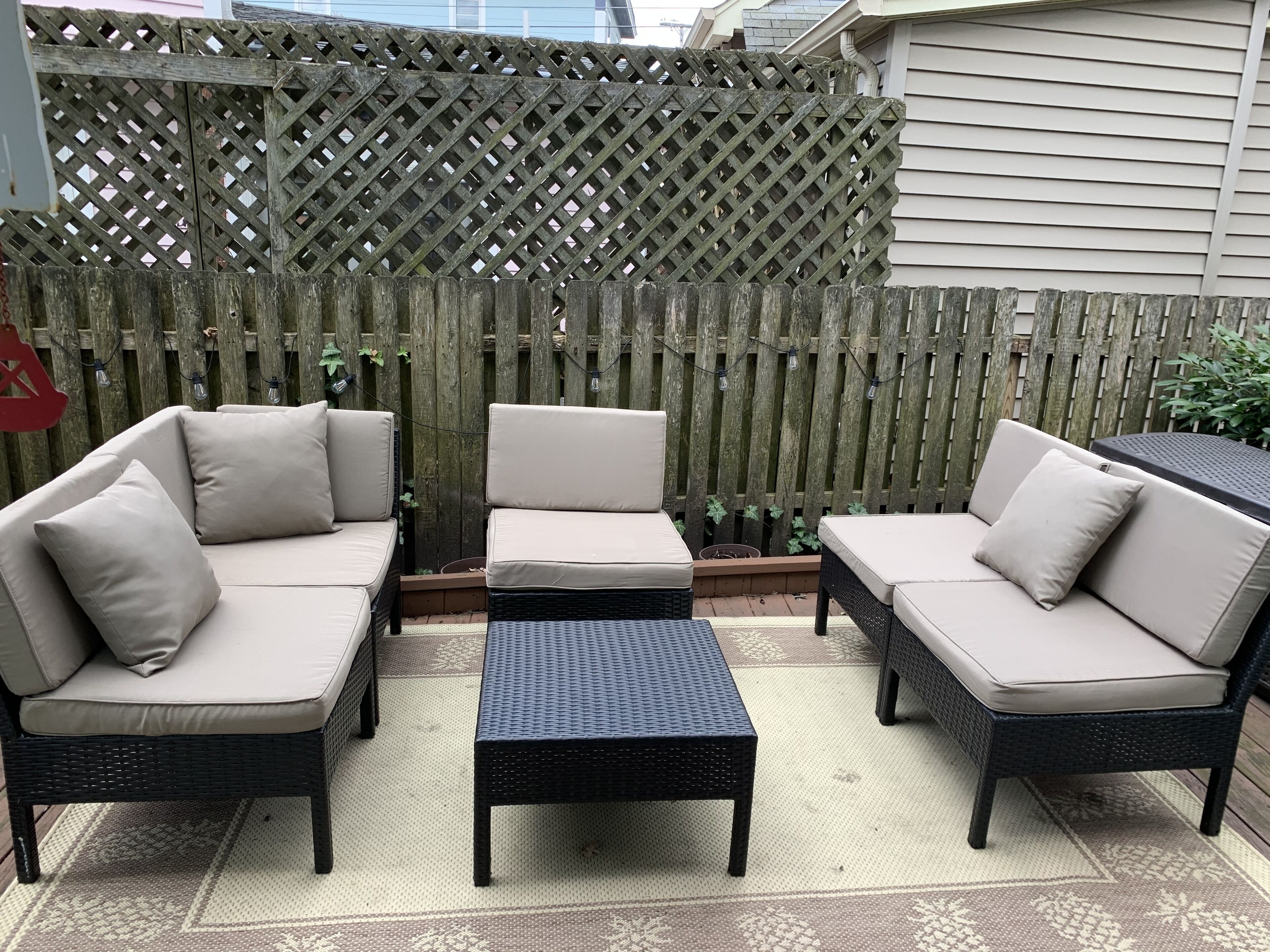 An outdoor patio with a wooden lattice fence, two beige sofas, a black woven coffee table, and a small table with a dark top.