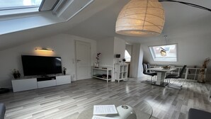 Una sala de estar moderna con un televisor de pantalla plana, una mesa de centro de vidrio con papeles, un comedor con mesa y sillas, y un ventanal con vistas.