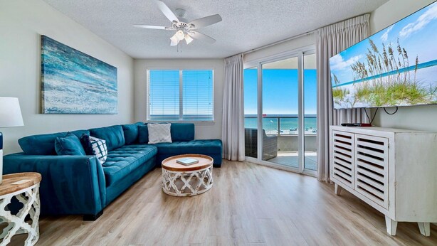 Un salon moderne avec un canapé bleu, un ventilateur de plafond, un grand téléviseur et une vue sur la plage.