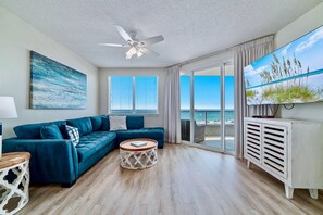 Un salon moderne avec un canapé bleu, un ventilateur de plafond, un grand téléviseur et une vue sur la plage.