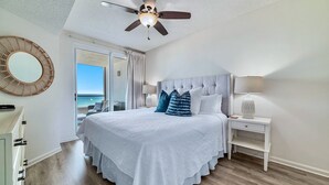 Une chambre à coucher comprenant un lit, une table de chevet, une lampe, un ventilateur de plafond, un grand miroir et une vue sur l’océan.