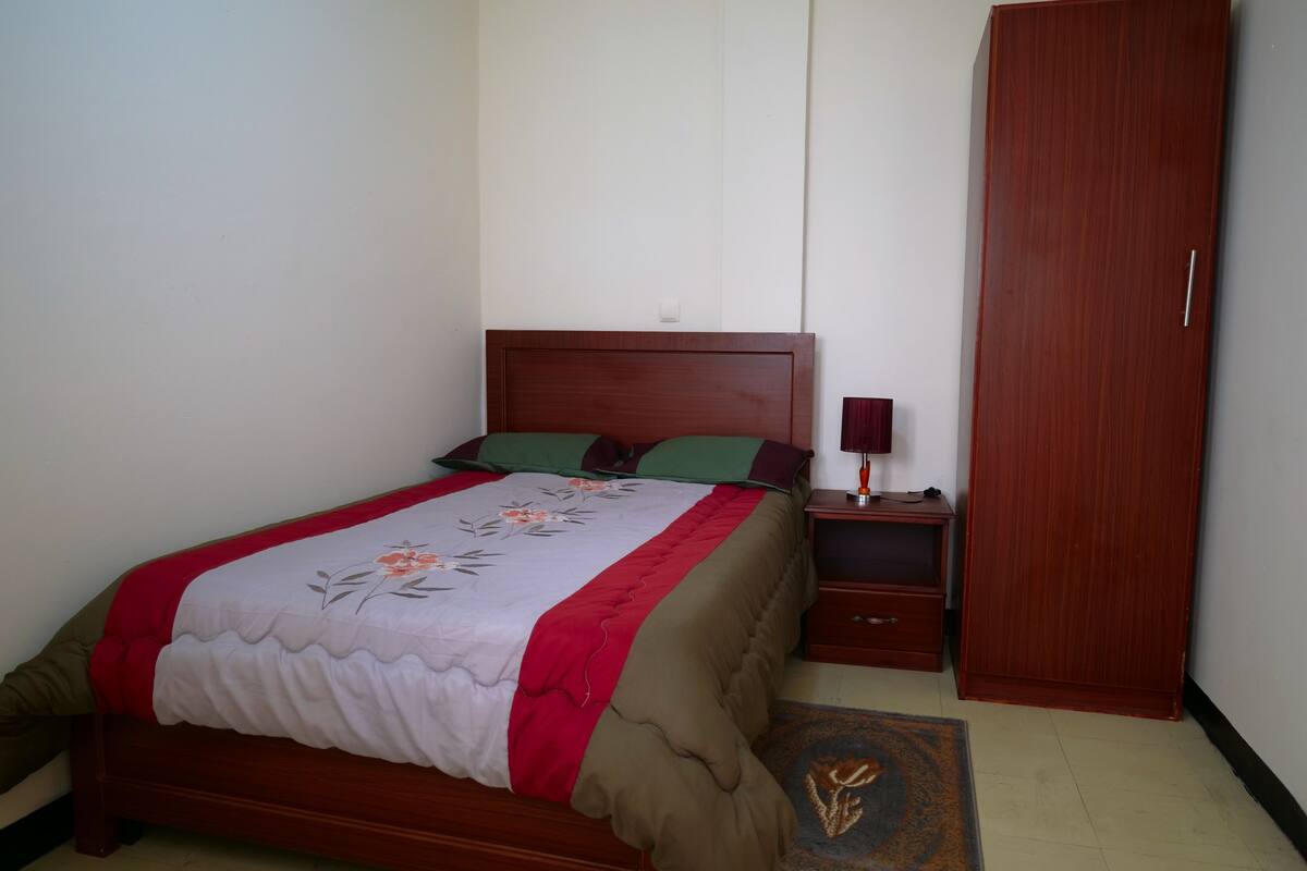 A neatly made bed with a floral bedspread, a bedside table with a lamp, and a wardrobe in a room with plain walls.