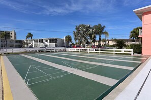 A green shuffleboard court with white lines and numbers, surrounded by a white fence, with buildings and palm trees in the background.
