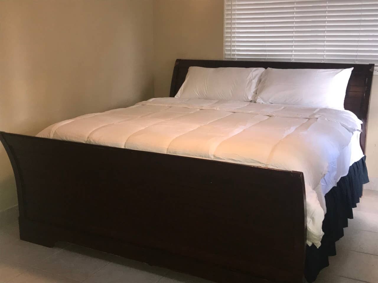 Een bed met een donker houten frame en wit beddengoed in een kamer met beige wanden en een raam met jaloezieën.