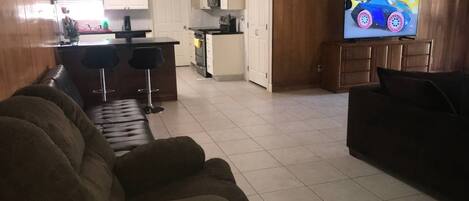 A living room with a dark brown sofa, a TV on the wall, and a kitchen in the background.