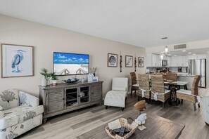 A living room with a television, framed artwork, and a dining area with wicker chairs. - 1 Step to the Beach! Beachfront Condo with 3 Sunset Decks, Sleeps 6 (Indian Rocks Beach)