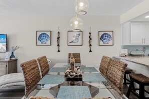 A modern dining area with a glass table, wicker chairs, and a hanging light fixture. - 1 Step to the Beach! Beachfront Condo with 3 Sunset Decks, Sleeps 6 (Indian Rocks Beach)