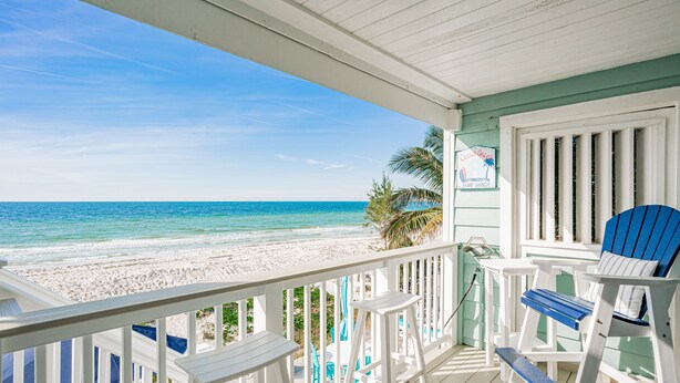 A beach view from a porch with white railings, a blue and white chair, and a sign for a surf shack. - 1 step and you are on the BEACH! Watch amazing sunsets from 3 decks! PremierHost (Indian Rocks Beach)