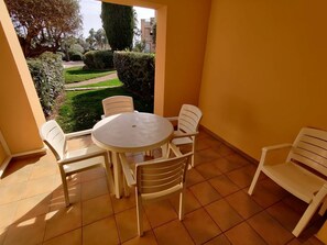 Eine kleine Terrasse mit rundem Tisch und vier Stühlen, ein Fliesenboden und Blick auf einen Garten sowie Gebäude im Hintergrund.