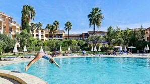 Piscina con sillas de descanso y sombrillas, rodeada de palmeras y edificios de hotel.