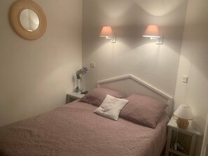 Ein Schlafzimmer mit einem Bett, zwei Wandlampen, einem runden Spiegel, einem Nachttisch mit einer Vase und einer Lampe sowie einer gemusterten Bettdecke.