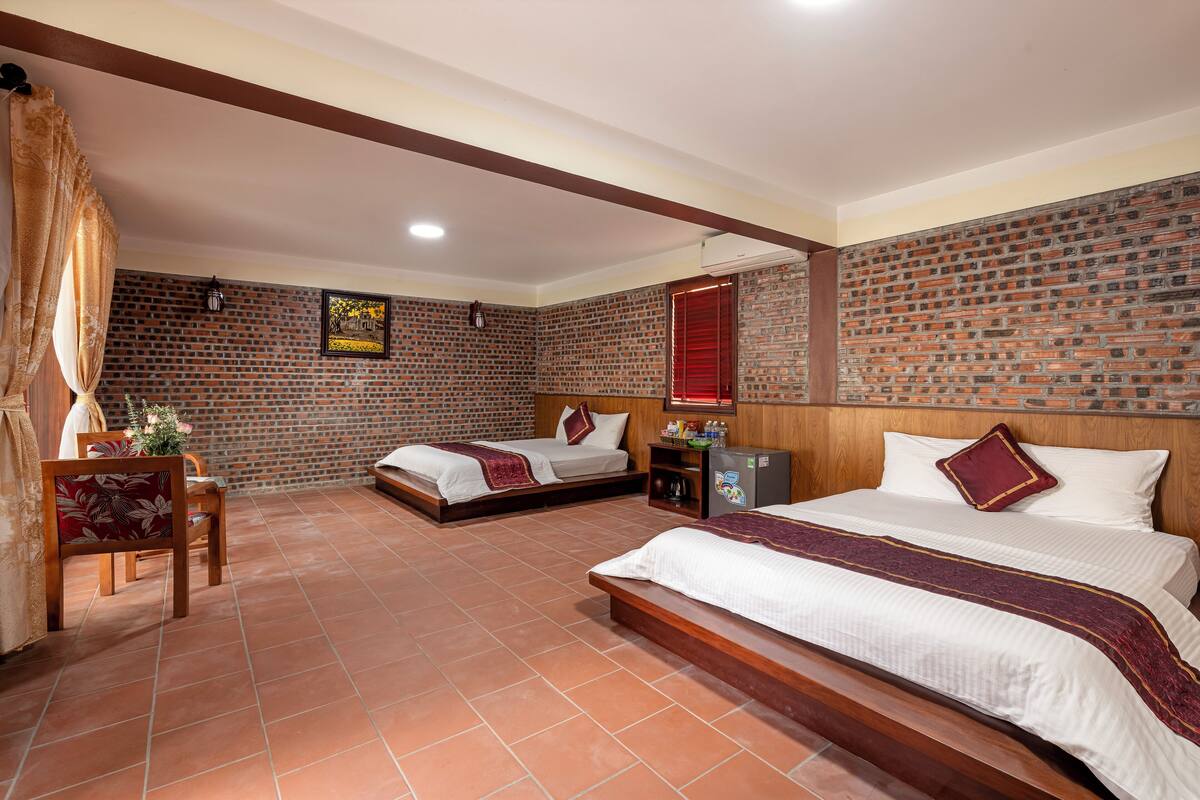 A hotel room with two beds, a wooden bench, a small table with a chair, a refrigerator, and a brick-patterned wall.