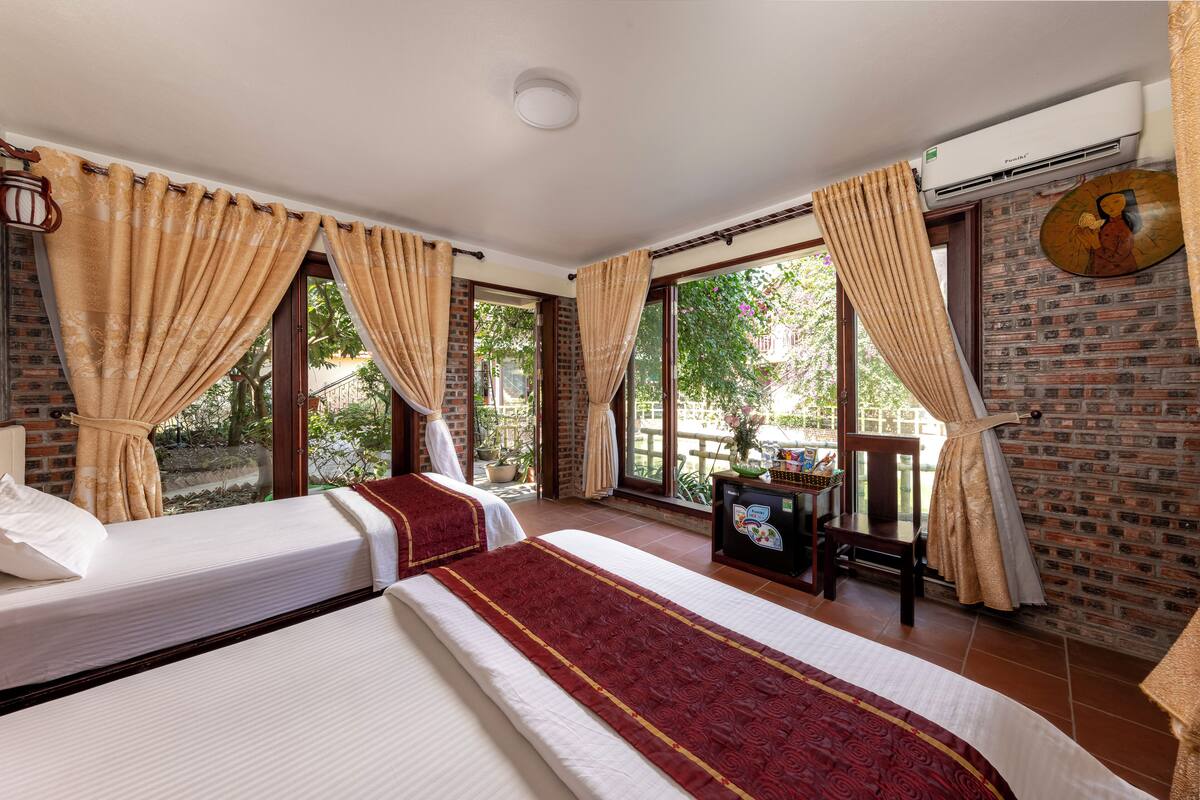 A hotel room with two beds, large windows with curtains, a brick wall, and a ceiling fan.