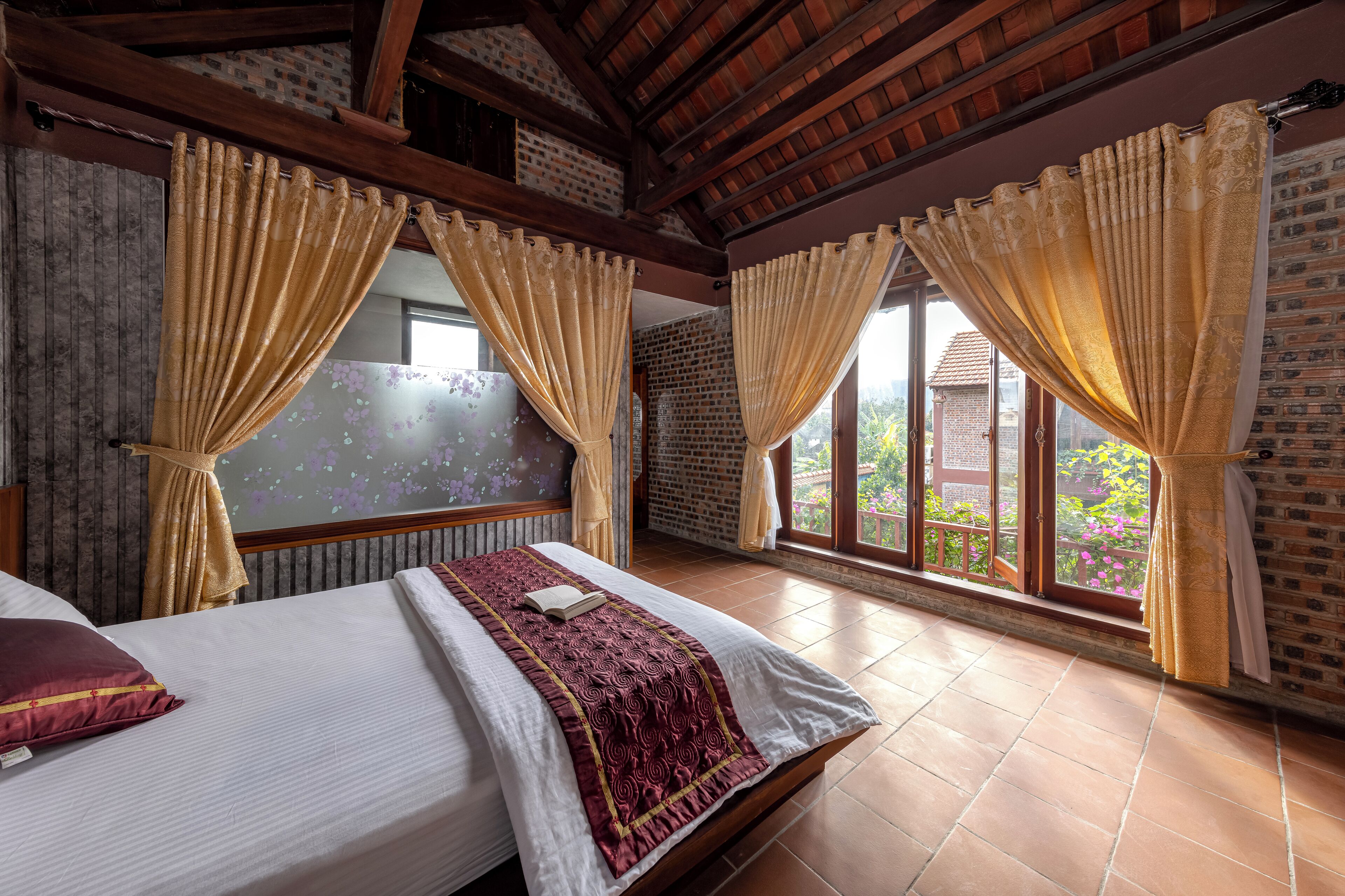 Quarto de hotel com uma cama grande, duas janelas com cortinas, uma parede de tijolos e piso de azulejos.