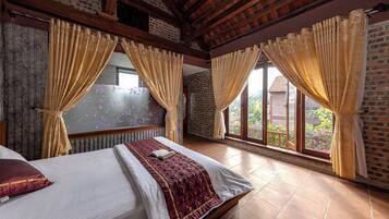 Quarto de hotel com uma cama grande, duas janelas com cortinas, uma parede de tijolos e piso de azulejos.