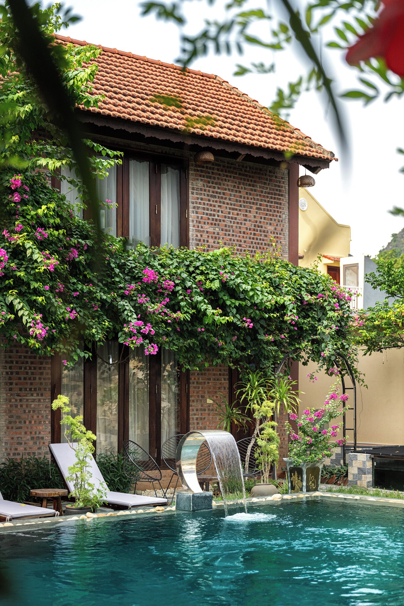 A pool area with a brick building, a pergola covered in flowering vines, and a water feature.