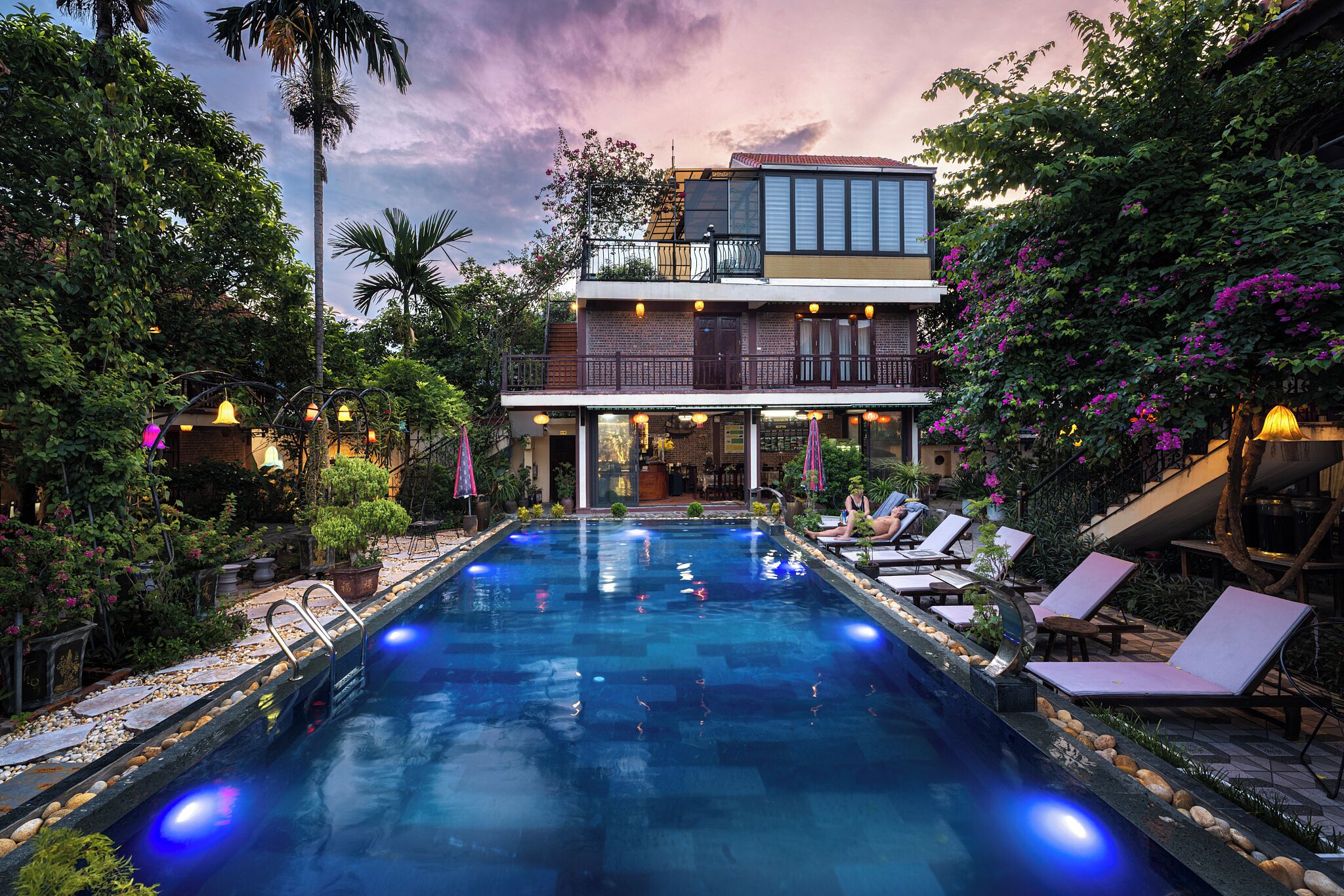 A swimming pool with lounge chairs, surrounded by lush greenery and a two-story building with a balcony.