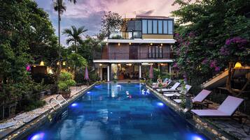 A swimming pool with lounge chairs, surrounded by lush greenery and a two-story building with a balcony.