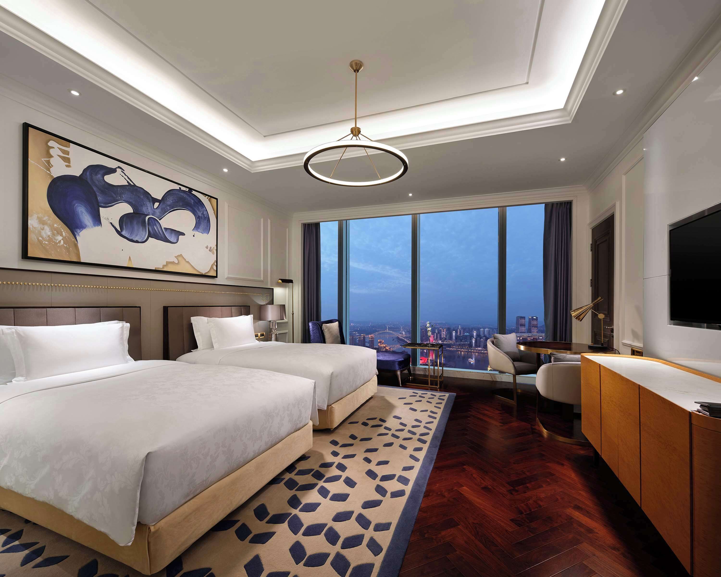 A hotel room with two beds, a desk, a chair, a large window with a city view, and a modern chandelier.