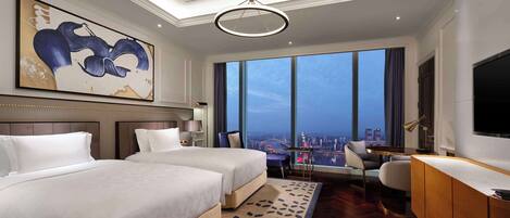 A hotel room with two beds, a desk, a chair, a large window with a city view, and a modern chandelier.
