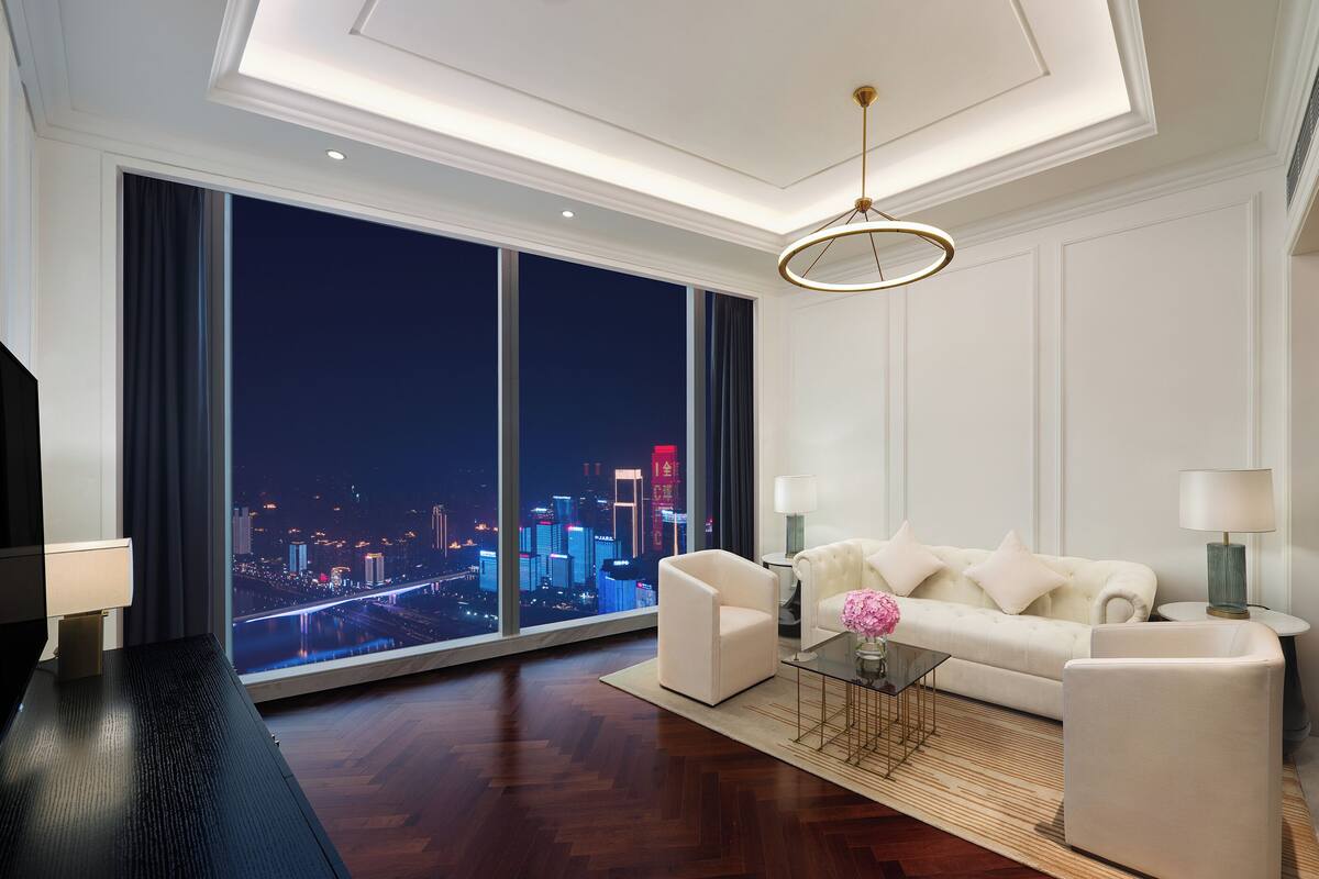 A modern hotel room with a large window offering a city view, a white sofa, a glass coffee table, and a minimalist decor style.
