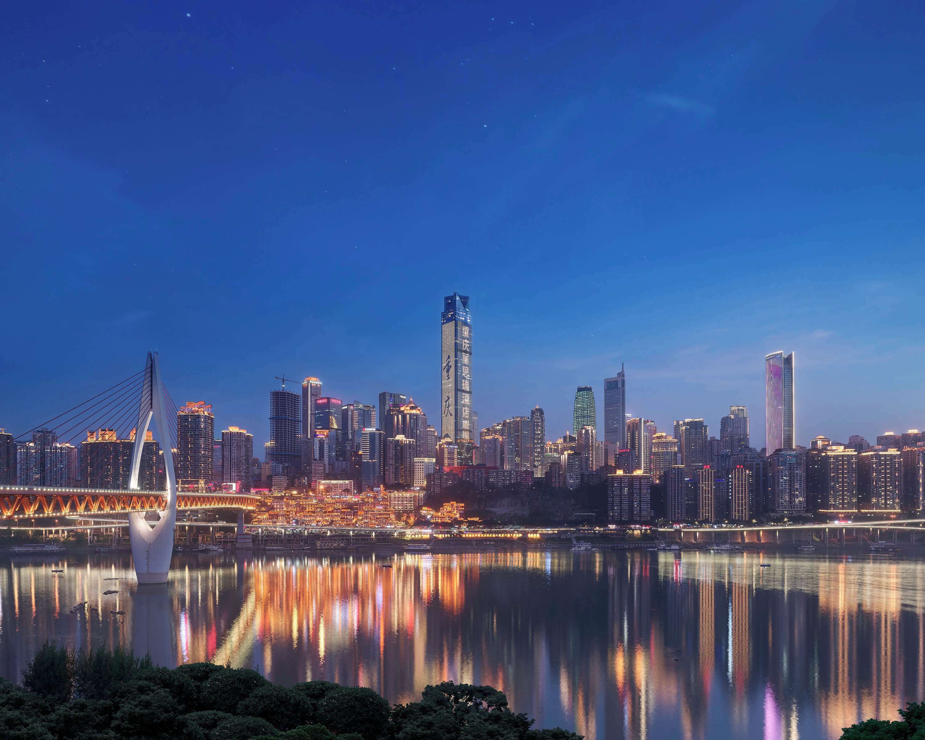 Un horizonte urbano al anochecer con edificios iluminados reflejándose en un cuerpo de agua tranquilo, un puente destacado y un rascacielos moderno.