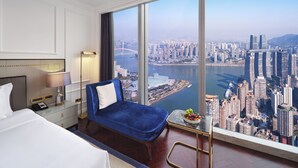 A hotel room with a large window offering a city view, a bed, a blue chair, a small table with fruit, and a lamp. - The Lixury Hotel -The Unbound Collection by Hyatt (Chongqing)