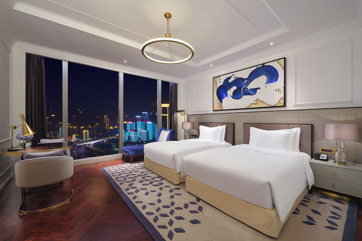 A hotel room with two beds, a city view, a large abstract painting, and a modern chandelier.