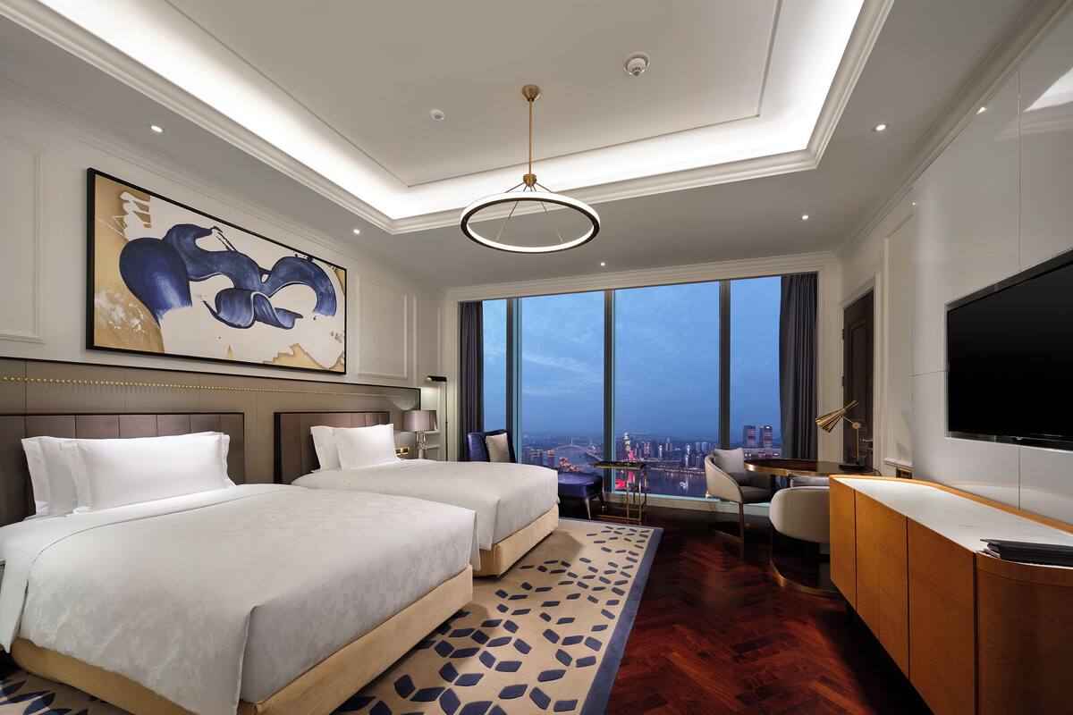 A hotel room with two beds, a desk, a TV, and a large window with a city view.