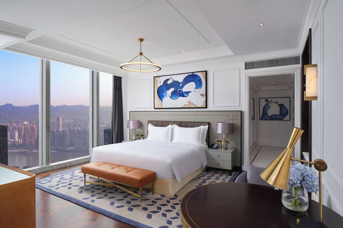 A modern hotel room with a large bed, a view of the city, and a round table with a vase of flowers.
