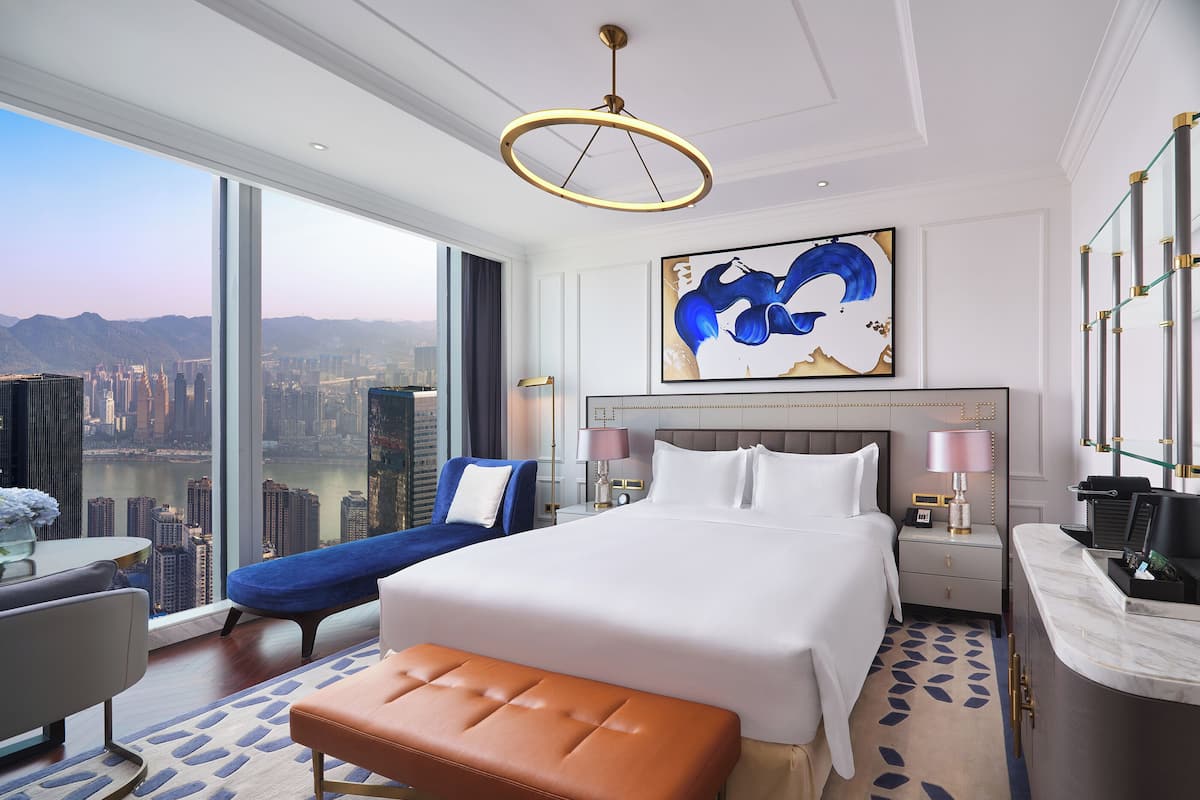 A modern hotel room with a large bed, a bench, a desk, and a view of the cityscape.