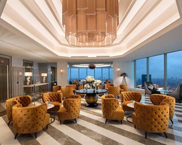 A spacious lobby with a central round table surrounded by yellow upholstered chairs, a modern chandelier, and large windows offering a city view.