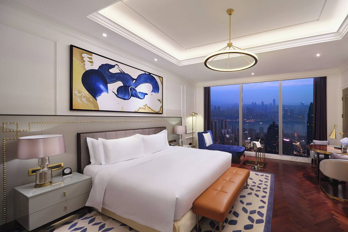 A modern hotel room with a large bed, a seating area, and a view of the cityscape.