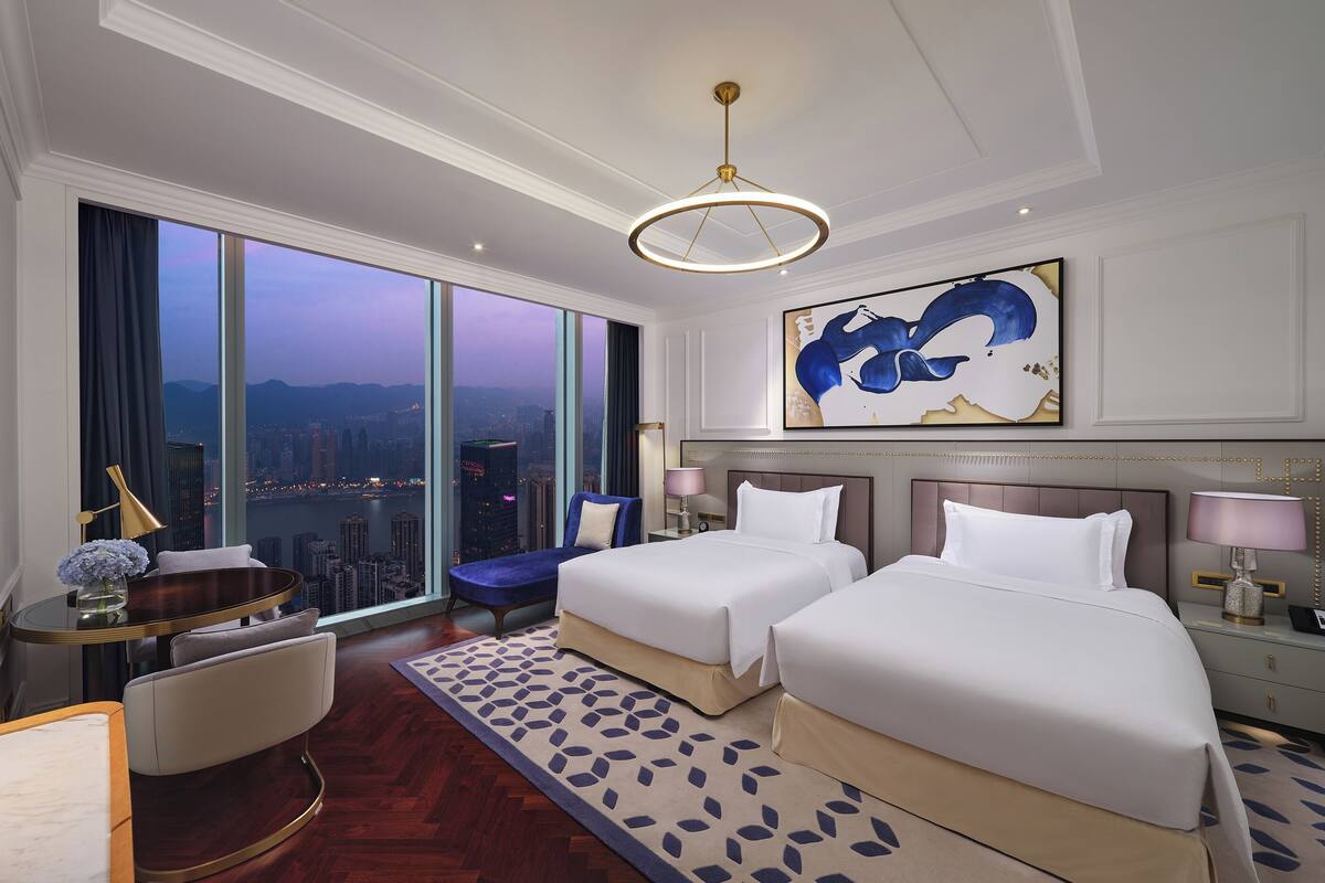 A hotel room with two beds, a desk, a chair, a large window with a city view, and a modern chandelier.