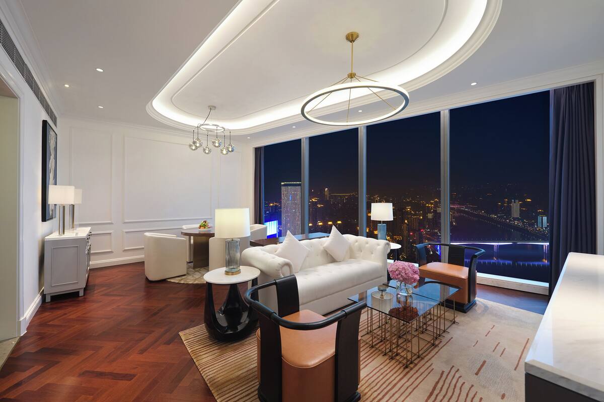 A modern hotel room with a large city view, a white sofa, a glass coffee table, and a round floor lamp.