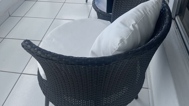 Un balcón con dos sillas de mimbre, una con cojín blanco y una pequeña planta en maceta.