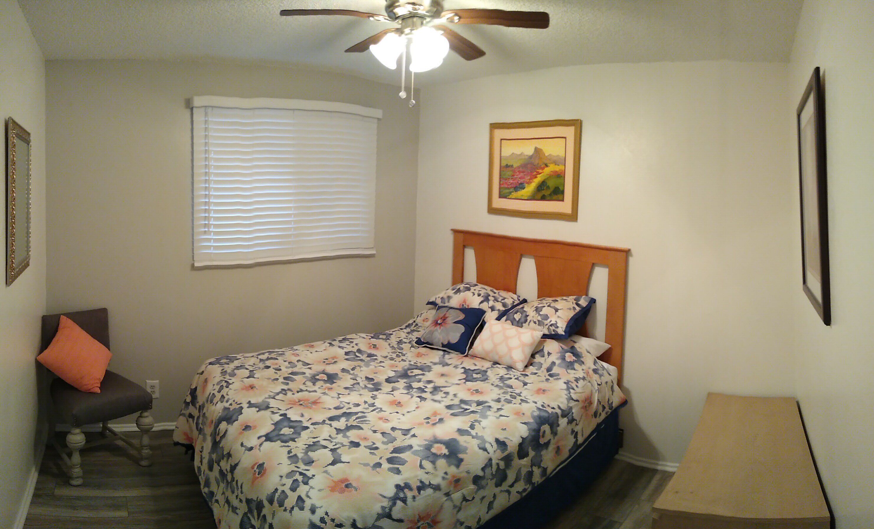 Um quarto com cama, ventoinha no teto, janela com persianas, quadro pendurado na parede e uma cadeira com almofada.