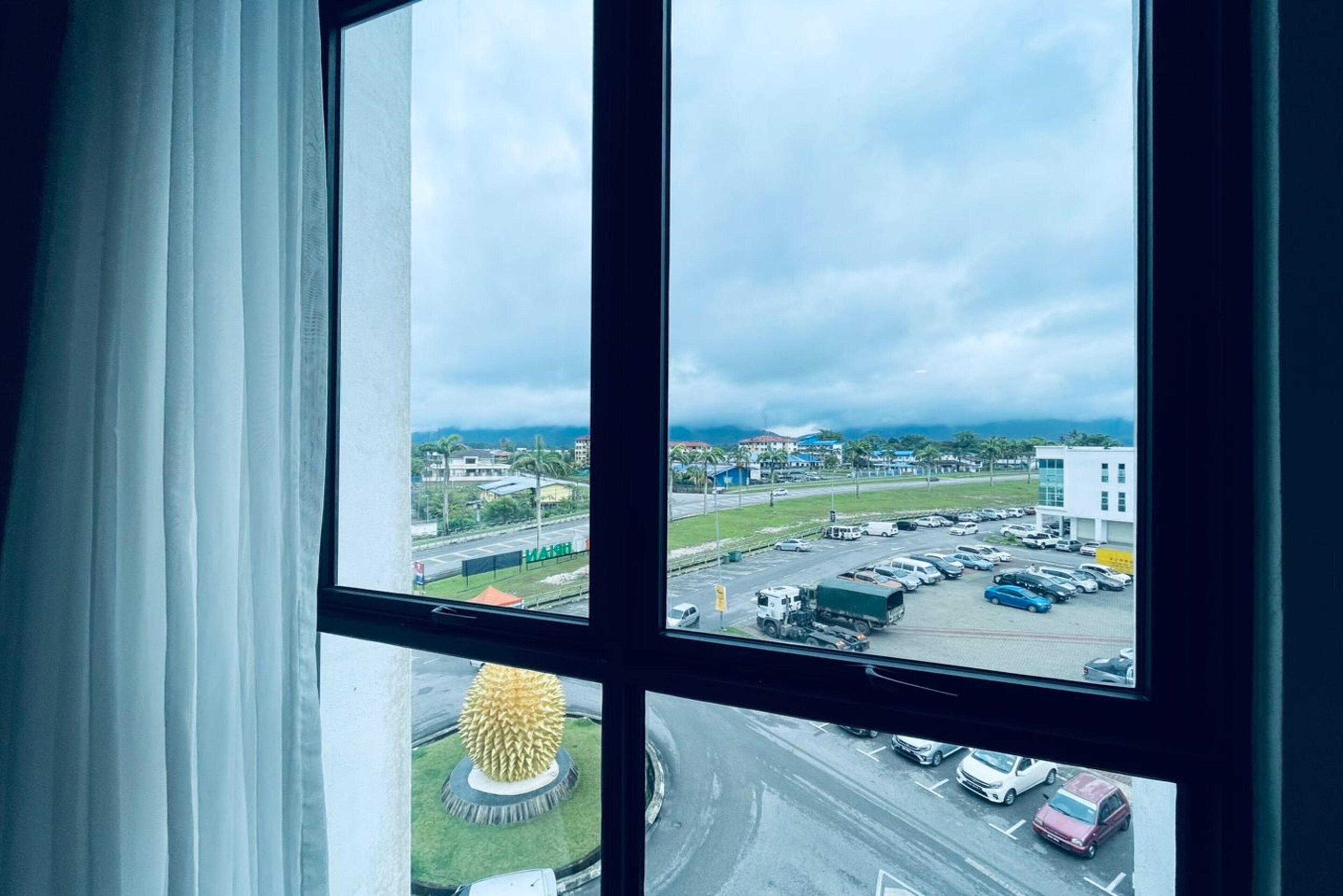 Vista desde una ventana con cortinas blancas, que da a un estacionamiento con varios coches y un edificio al fondo.