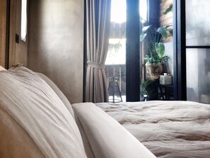 Ein Schlafzimmer mit einem großen Bett, Blick ins Freie durch ein Fenster und einem Topf mit einer Pflanze auf einem Tisch.