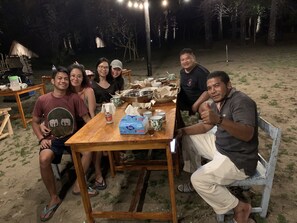 A group of people seated around a wooden table outdoors at night, with a thatched-roof structure and trees in the background.