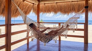 En hängmatta under en takkonstruktion av halmtak, med utsikt över stranden och havet.