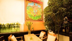Un espace extérieur accueillant, avec des tables et des chaises en bois, une fresque murale décorative et un grand arbre qui offre de l’ombre.