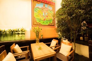 A cozy outdoor seating area with wooden tables and chairs, a decorative wall mural, and a large tree providing shade.