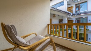 A balcony with a wooden chair, a white textured wall, and a view of a building with a tiled roof and windows.