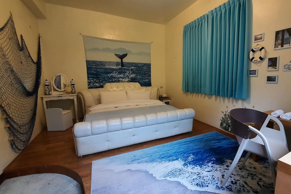A bedroom with a bed, a desk, a chair, a large window with curtains, and a wall with a whale tail painting.