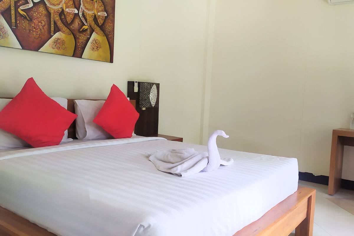 A neatly made bed with red and white pillows, a swan-shaped towel, and a painting on the wall.