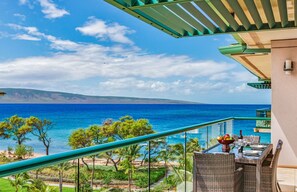 A balcony with a view of the ocean, wicker furniture, and a table with a fruit bowl and wine bottle.